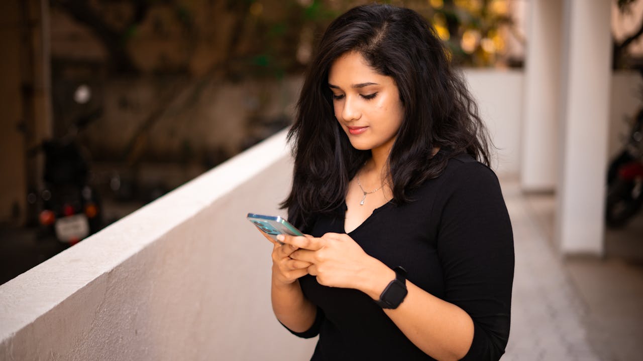 A young woman stands on a terrace, focused on her smartphone. Warm outdoor setting with casual attire.