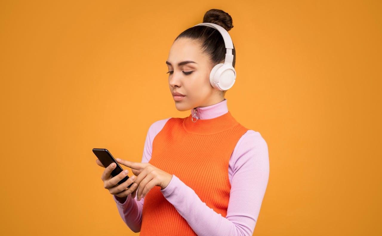 Woman with white headphones using smartphone against an orange background.