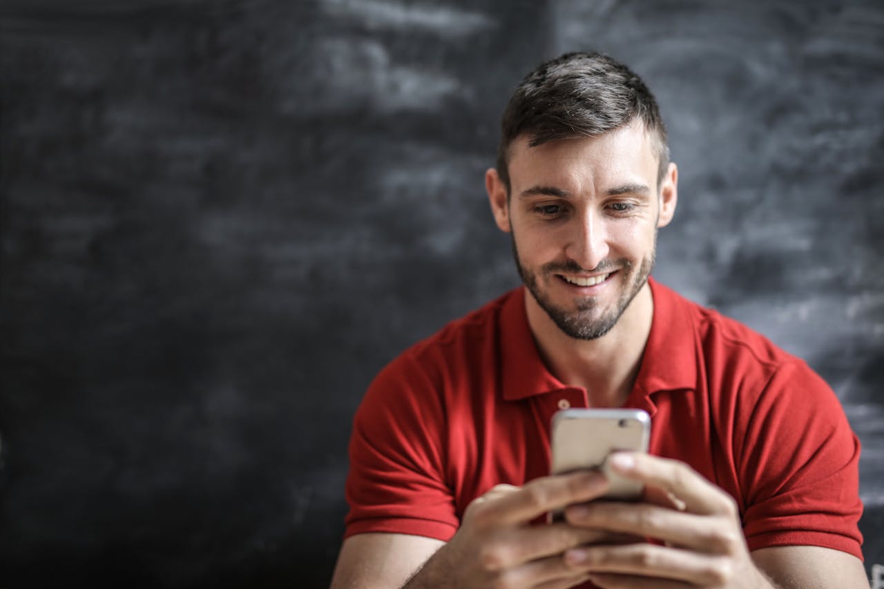 A man in a red polo shirt happily using his smartphone indoors against a dark background.