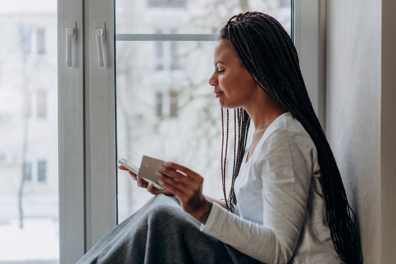 Side view of a woman sitting by a window, shopping online with a smartphone and credit card, enjoying leisure time indoors.