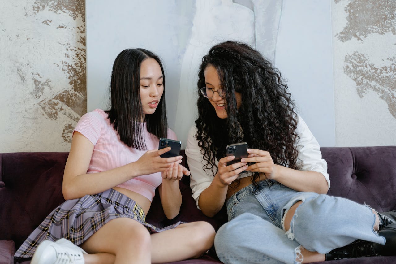 Two fashionable women sitting on a couch, engaged with their smartphones, sharing a moment.