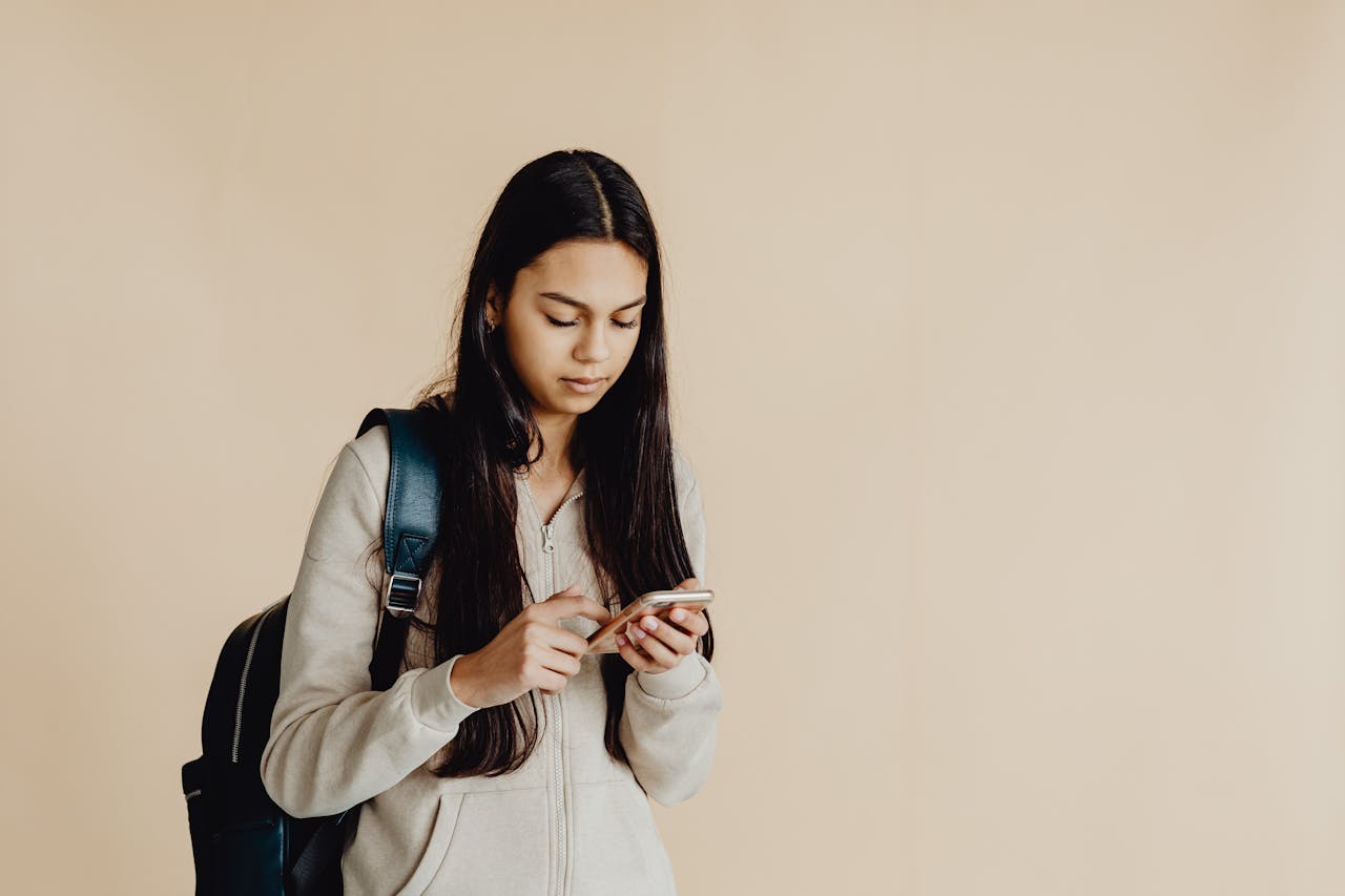 Teenage girl with backpack using a smartphone in a minimalist indoor setting, suited for educational themes.