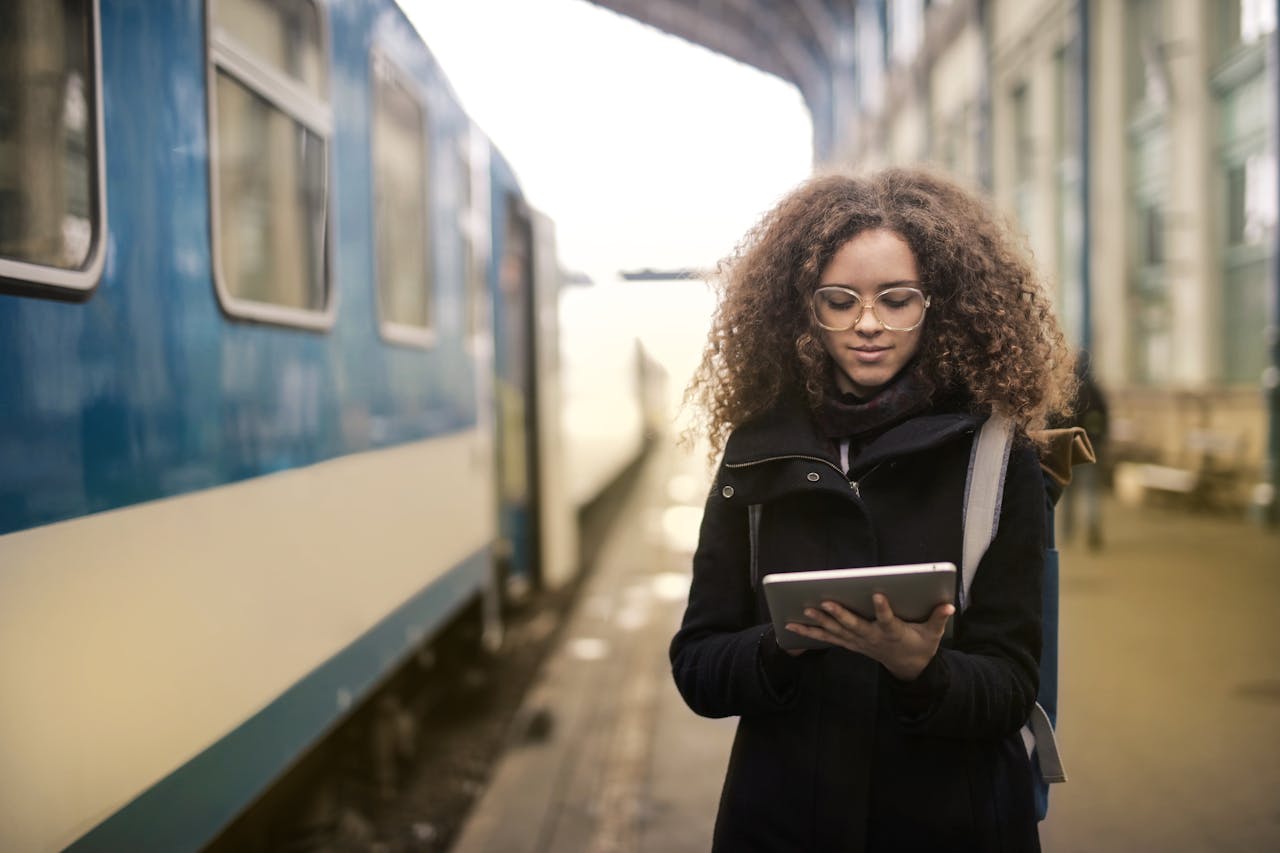A young woman with curly hair in a coat using a tablet at a train station platform.