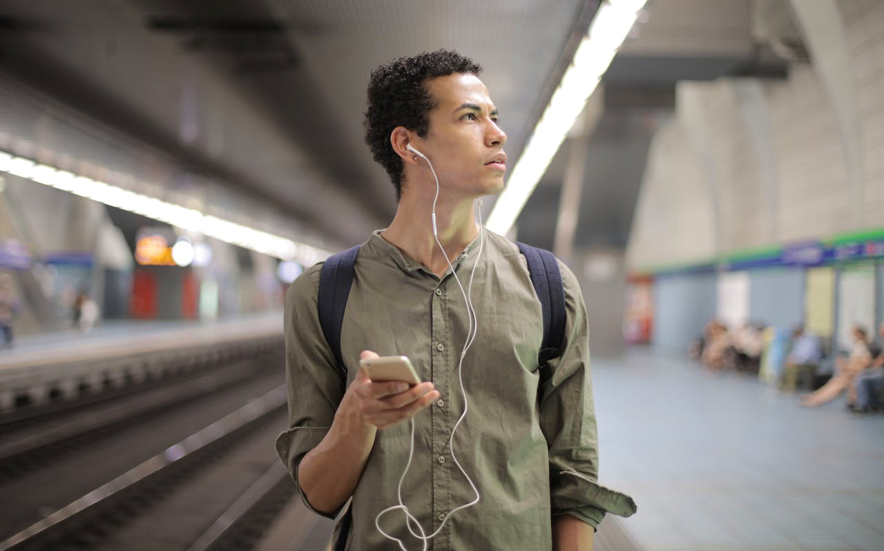 Portrait of a young man with earbuds waiting at a subway station holding a smartphone.
