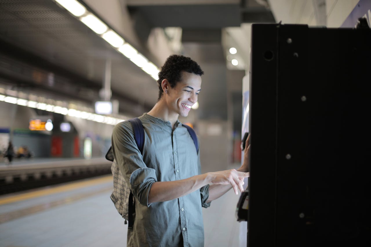 Joyful young African American male in casual clothes with backpack focusing and interacting with vending machine at underground station against blurred railway platform