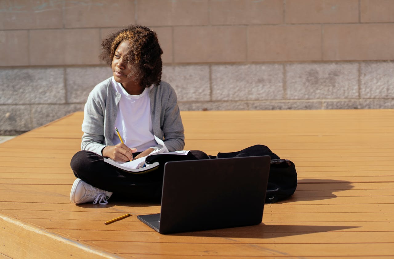 Contemplative African American schoolkid with copybook and netbook sitting with crossed legs on wooden platform while looking away in sunlight