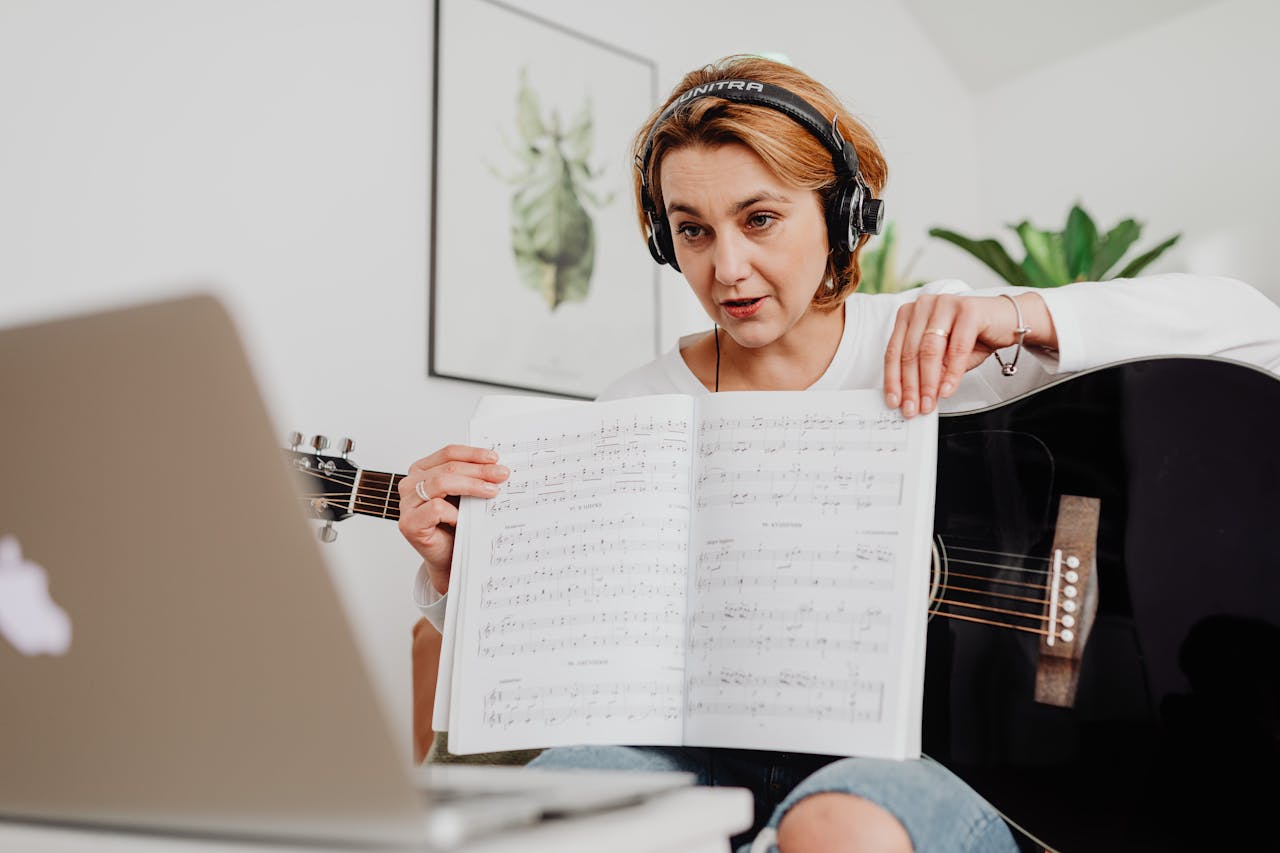 Woman teaching guitar online, presenting sheet music via video call with laptop setup.